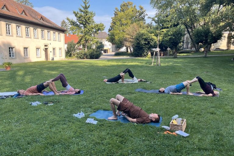 Yoga-Retreats - gemeinsame Auszeit Eine Gruppe von Personen macht gemeinsam Yoga-Übungen auf einer Wiese. Im Hintergrund Bäume und Gebäude.